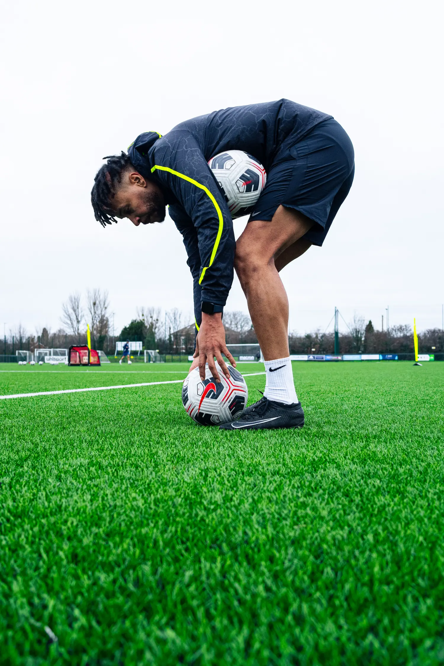 A football player in black athletic gear bends down on a lush green field, holding two soccer balls.