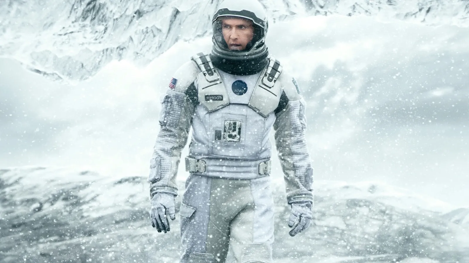An astronaut in a white spacesuit stands in a snowy, mountainous landscape, with frosty air and snowflakes visible, evoking a sense of isolation and exploration.