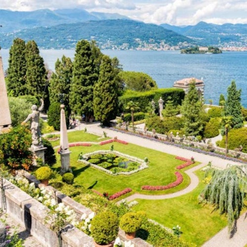 A beautifully landscaped lakeside garden with statues, well-maintained hedges, paths, and vibrant flower beds, set against mountains.