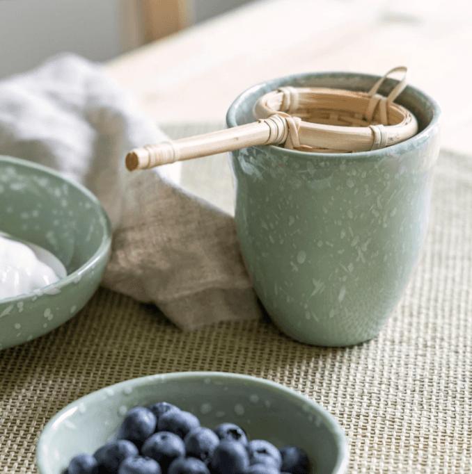 Mug en céramique mouchetée vert contenant des ustensiles en bambou, accompagné d'un bol de myrtilles, sur set de table en rotin
