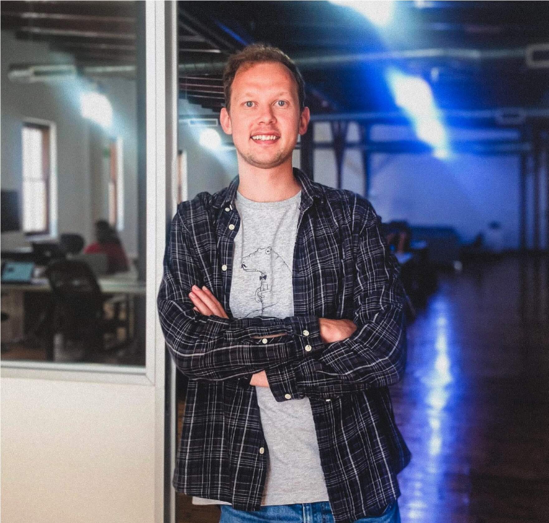 Smiling man with arms crossed, wearing a plaid shirt over a t-shirt, standing indoors.