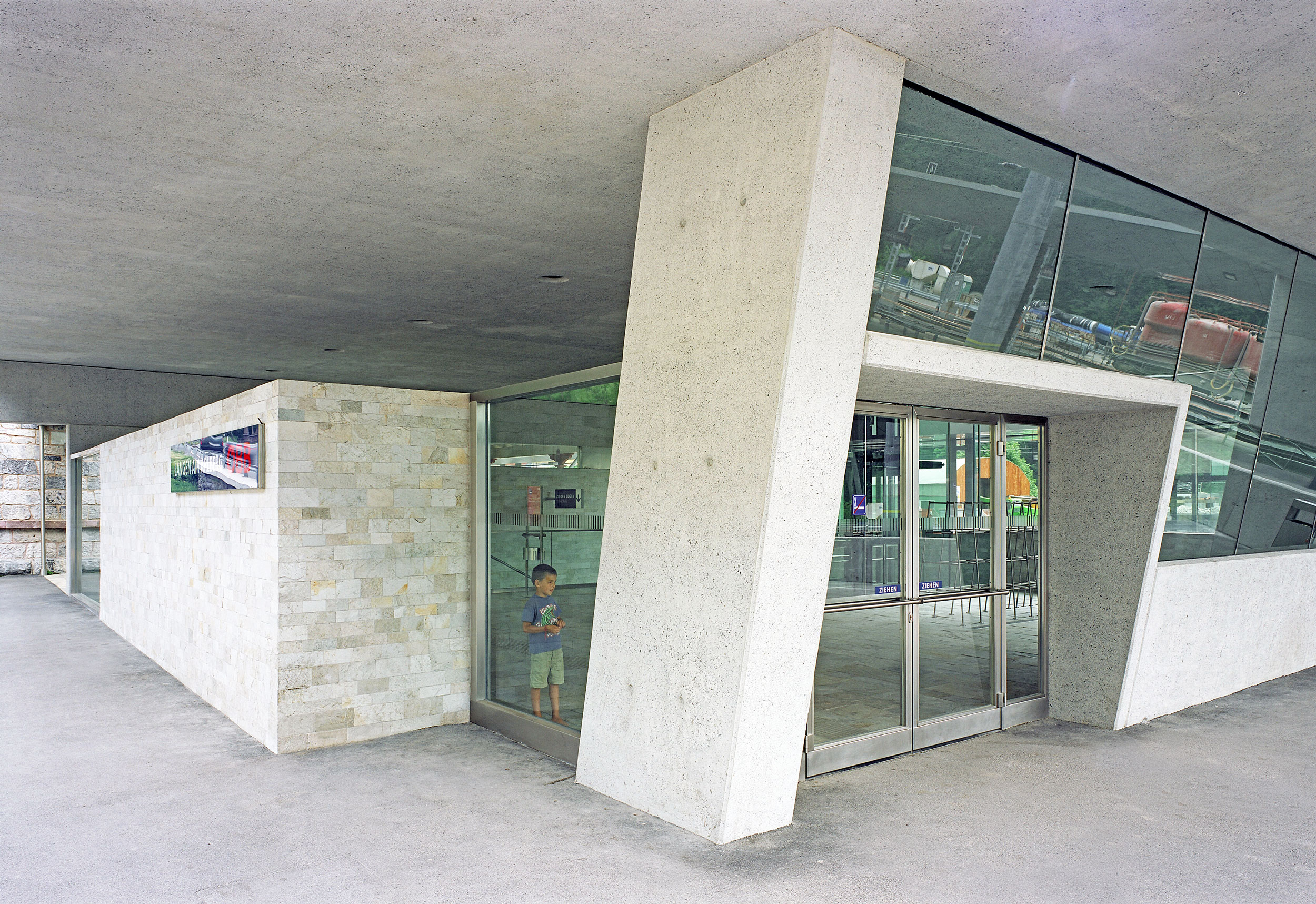 Gebäudeeingang mit geneigter Betonstütze, Glasfassade und Glastüren, innen sichtbare Person hinter der Tür.