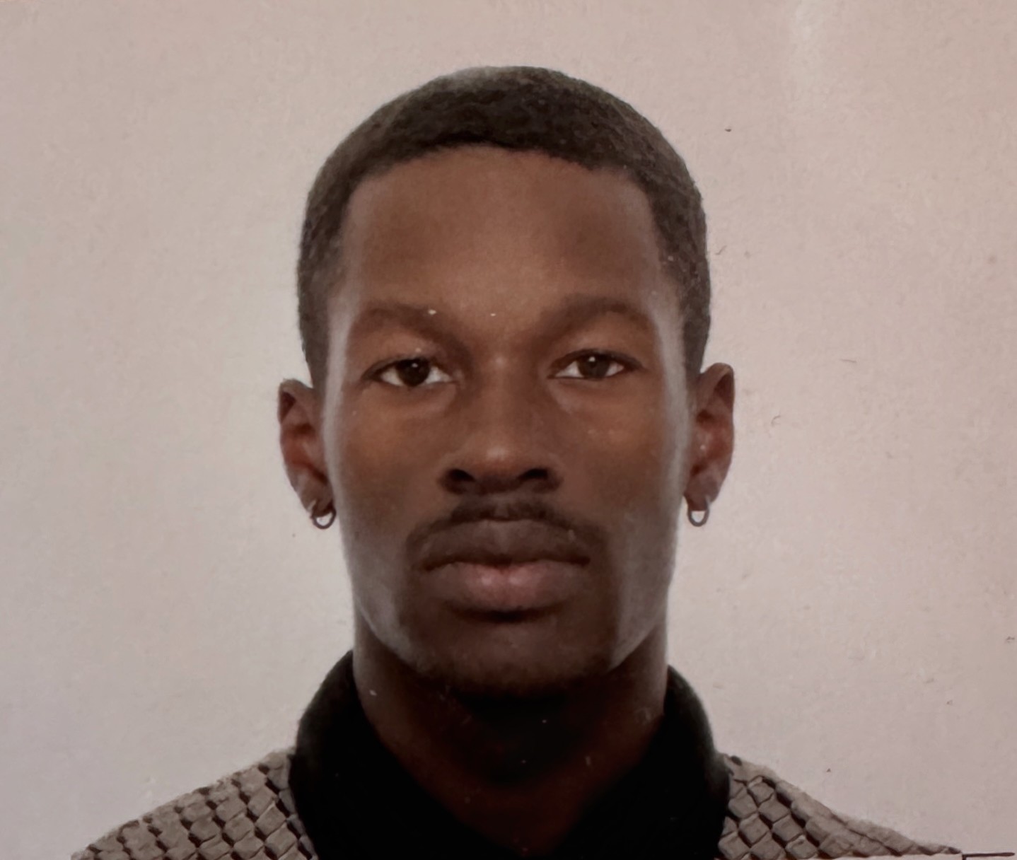 The passport photo of a twenty-something year old African-American male wearing a neutral expression and a shirt with a geometric pattern and dark collar, against a plain, softly lit background that looks like every DMV wall in America.