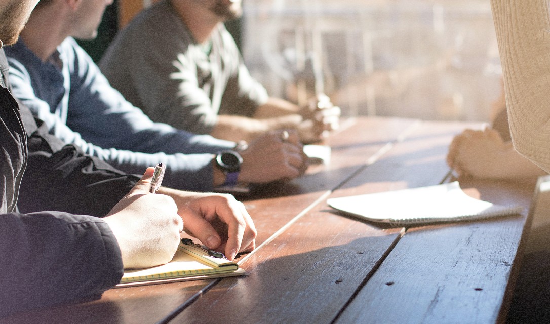 Vue d'une table de réunion avec plusieurs personnes en train de prendre des notes, des ordinateurs portables ouverts devant elles.