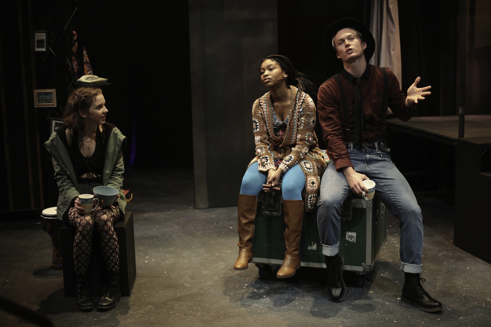 Three performers sit on road cases during a casual conversation. One performer in a green jacket watches as another, wearing a patterned cardigan and blue tights, listens to a performer in a hat who gestures animatedly with a mug in hand.