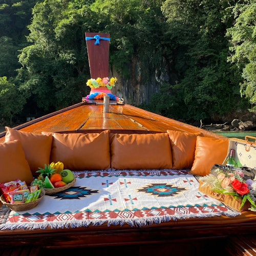 Fruit and snacks on a decorated blanket on a boat's deck, surrounded by brown cushions, with lush green trees in the background.