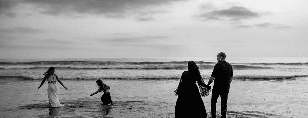 A black and white beach photograph at dusk. Two adults stand hand-in-hand at the water's edge, their backs to the camera, watching two children play in the surf. The overcast sky and rolling waves frame a quiet moment of connection.
