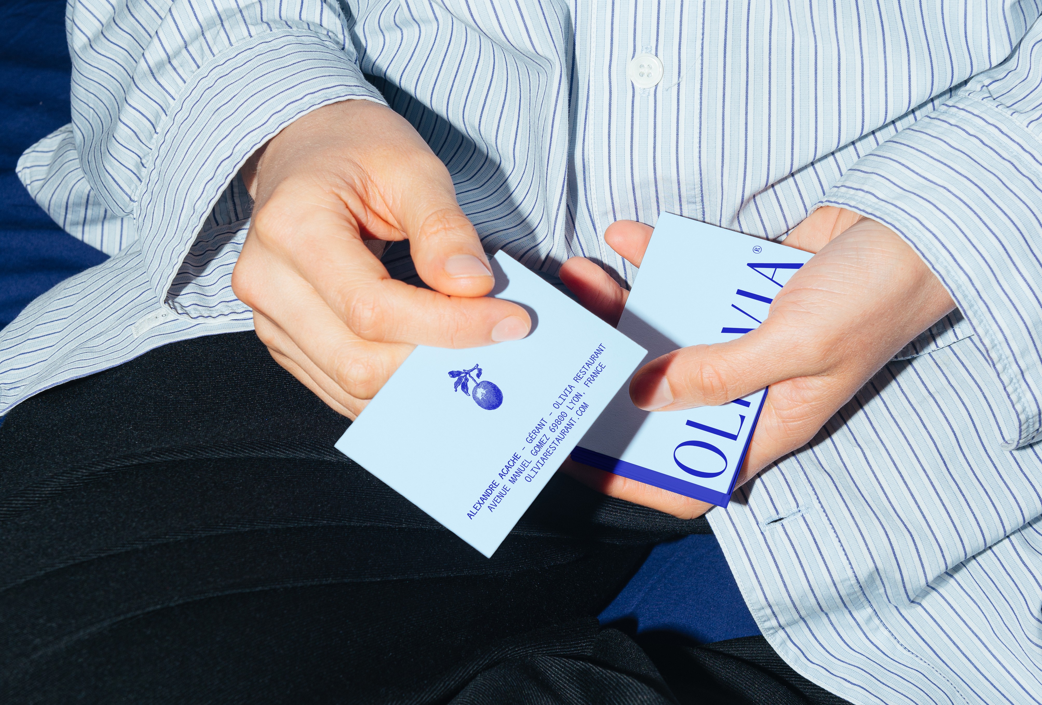 Cartes de visite du restaurant OLI•VIA tenues en main, recto bleu clair et verso bleu intense, design minimal et raffiné pour cuisine méditerranéenne.