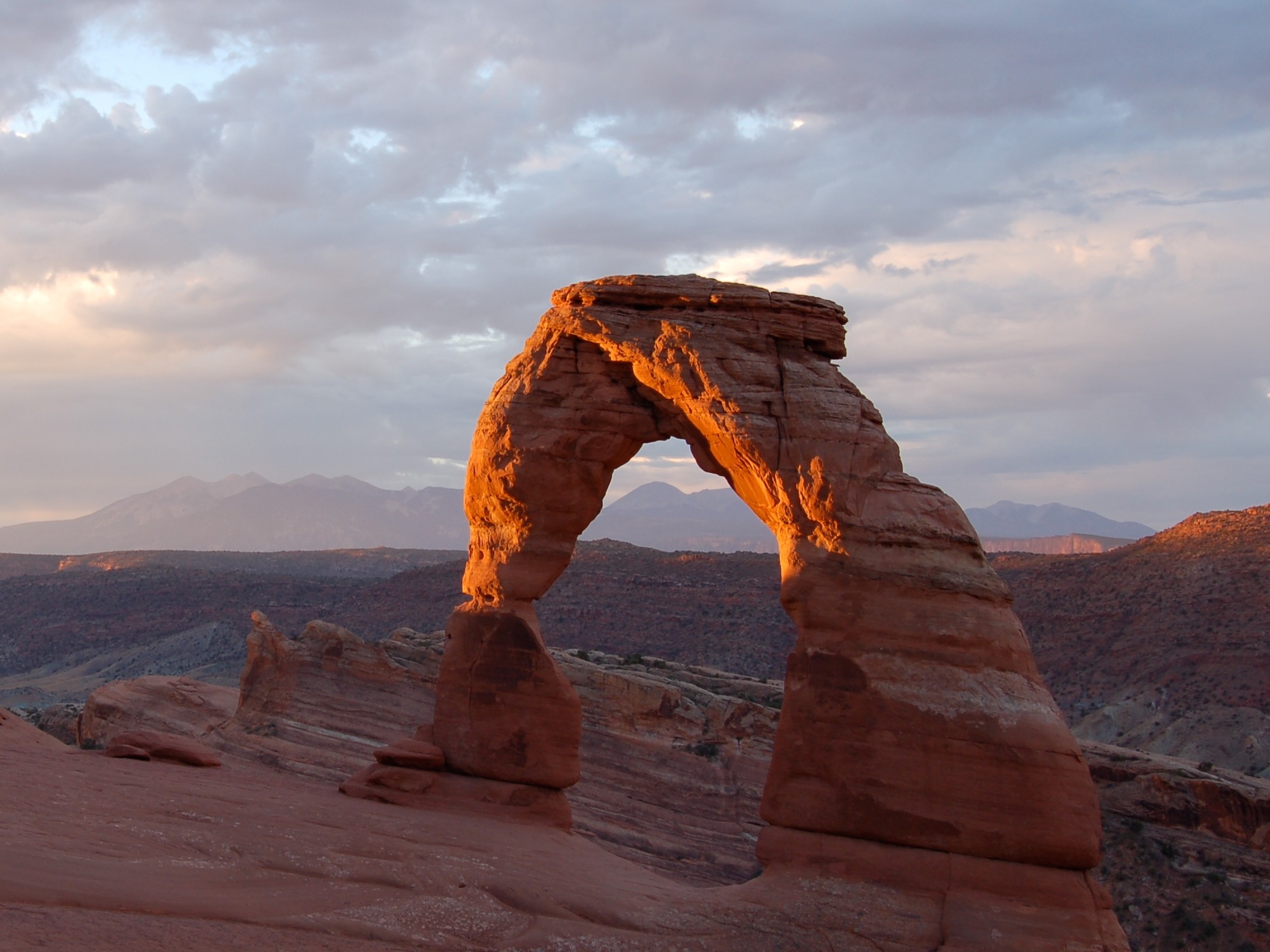 Delicate Arch in Arches National Park lit orange by low evening sun, standing free on slickrock with the hazy La Sal Mountains behind it.