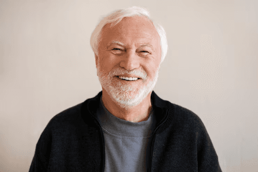 A smiling senior man with white hair and a beard, wearing a blue shirt and black jacket.