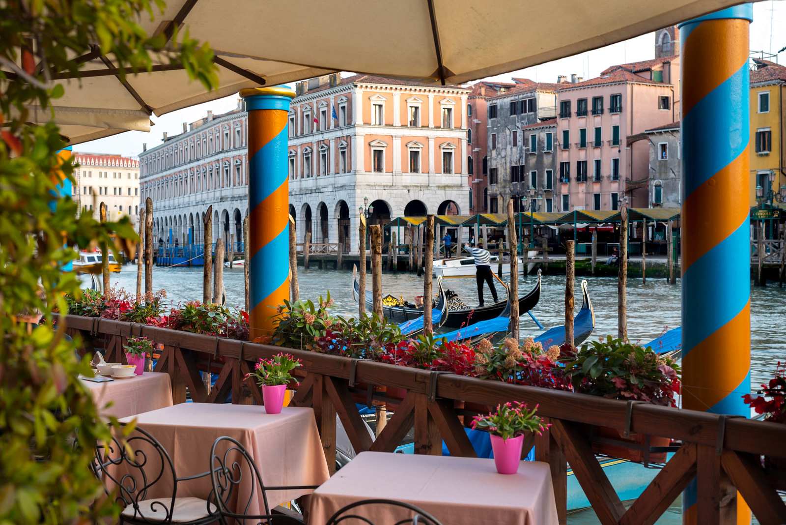 A waterfront cafe with white tables and pink flowers overlooks a canal lined with colorful buildings in Venice.