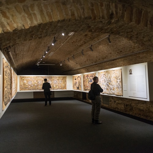 Two people view intricate paintings in a dimly lit gallery with a brick arched ceiling.