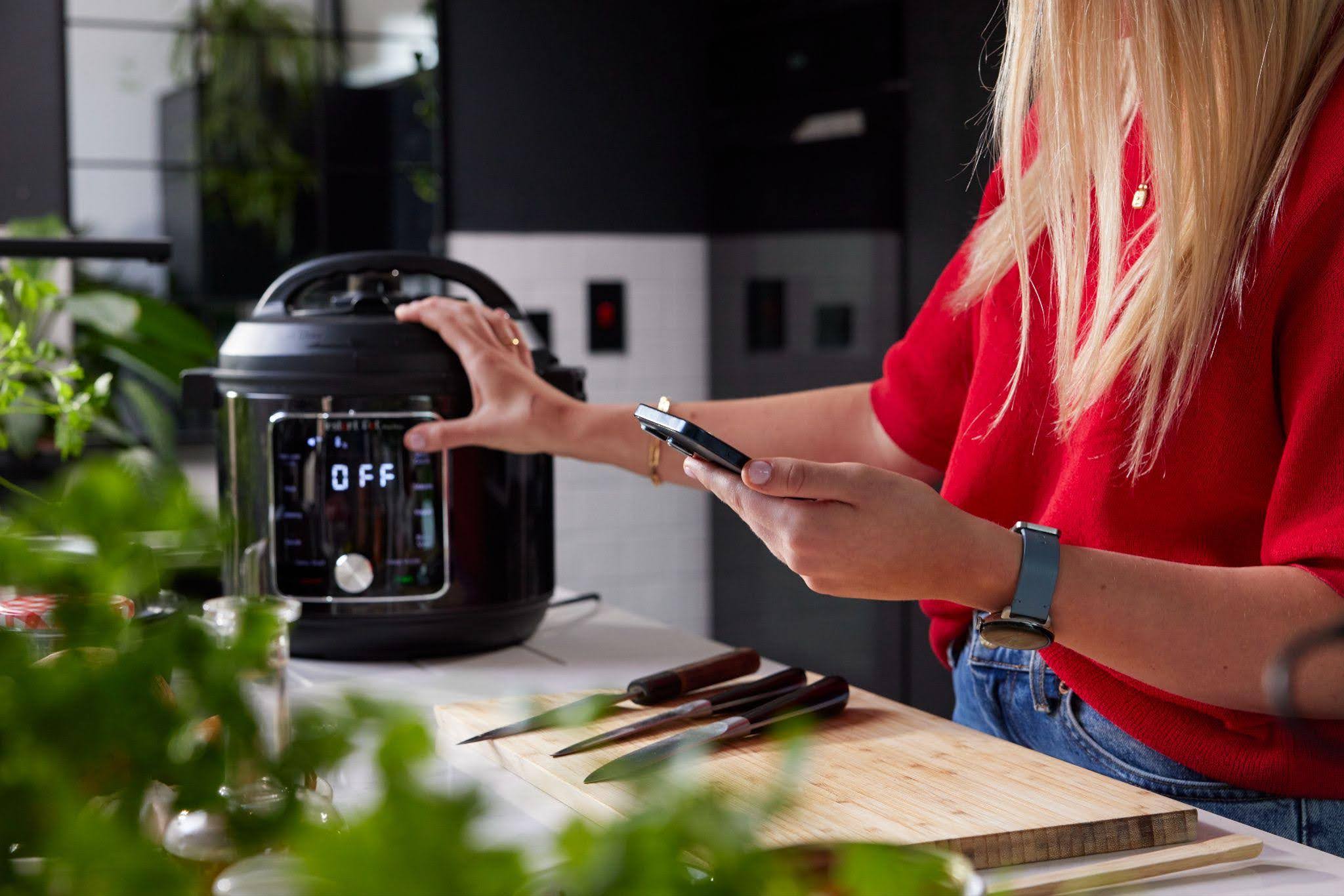 woman in a kitchen, holding a phone and pressing a button on an electric pressure cooker