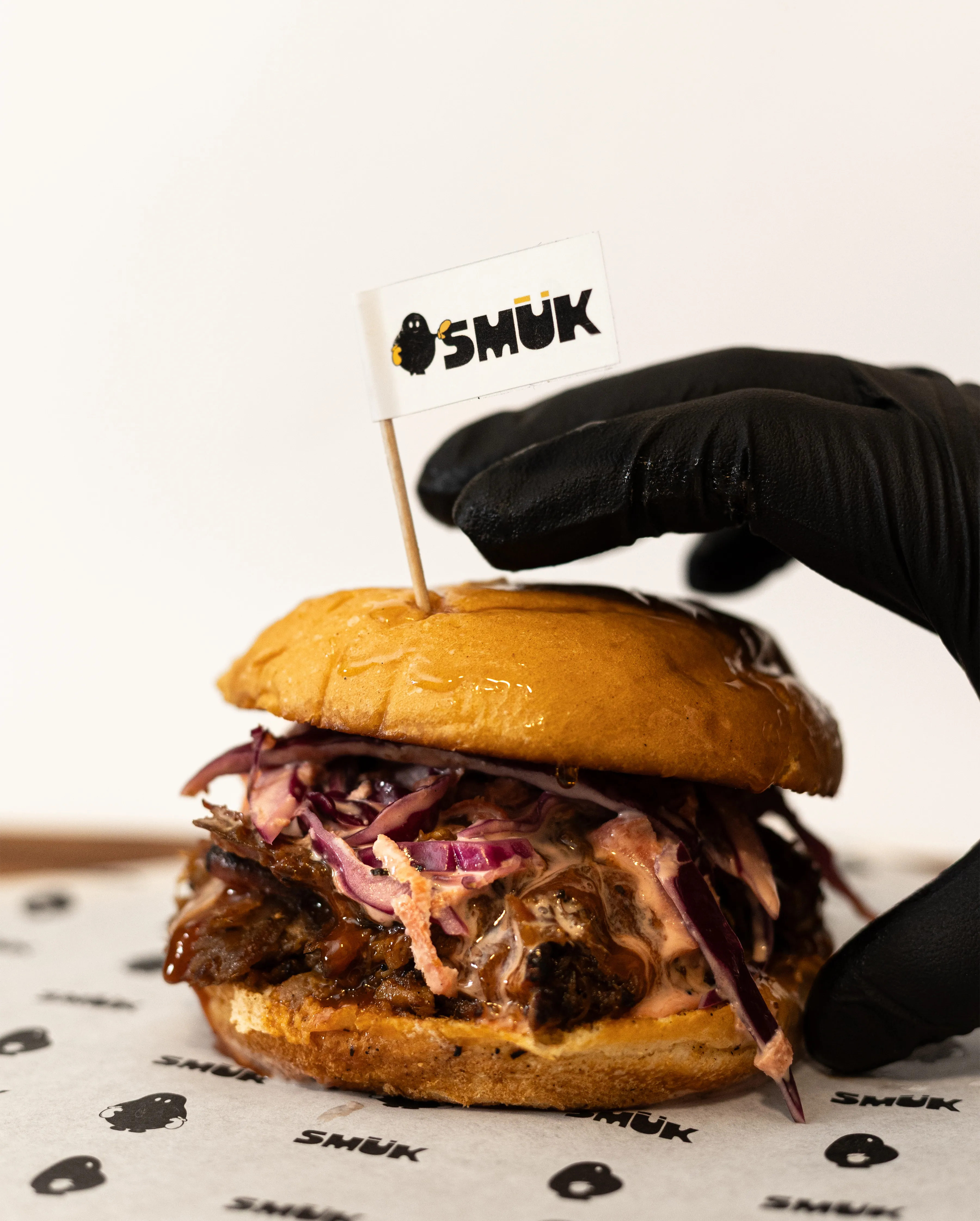 Mano con guante negro sosteniendo hamburguesa Smük con bandera