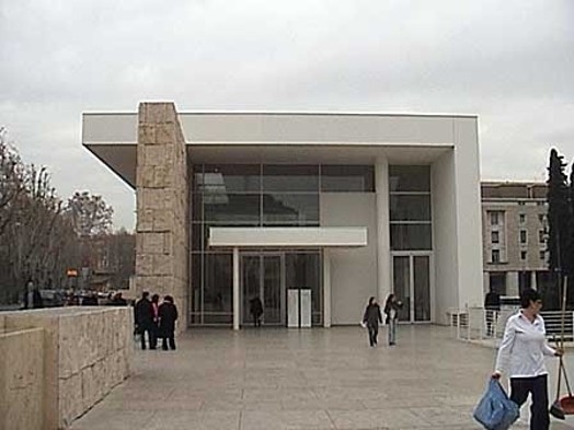 A modern cubical building with glass windows and a stone wall near it.