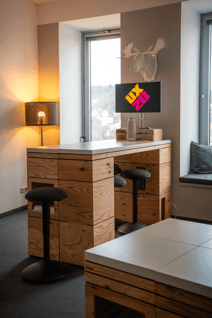 Moderner Arbeitsplatz mit hohem Holztisch, drei schwarzen Hockern, Fensterbank mit Kissen und Wandlampe in einem hellen Raum