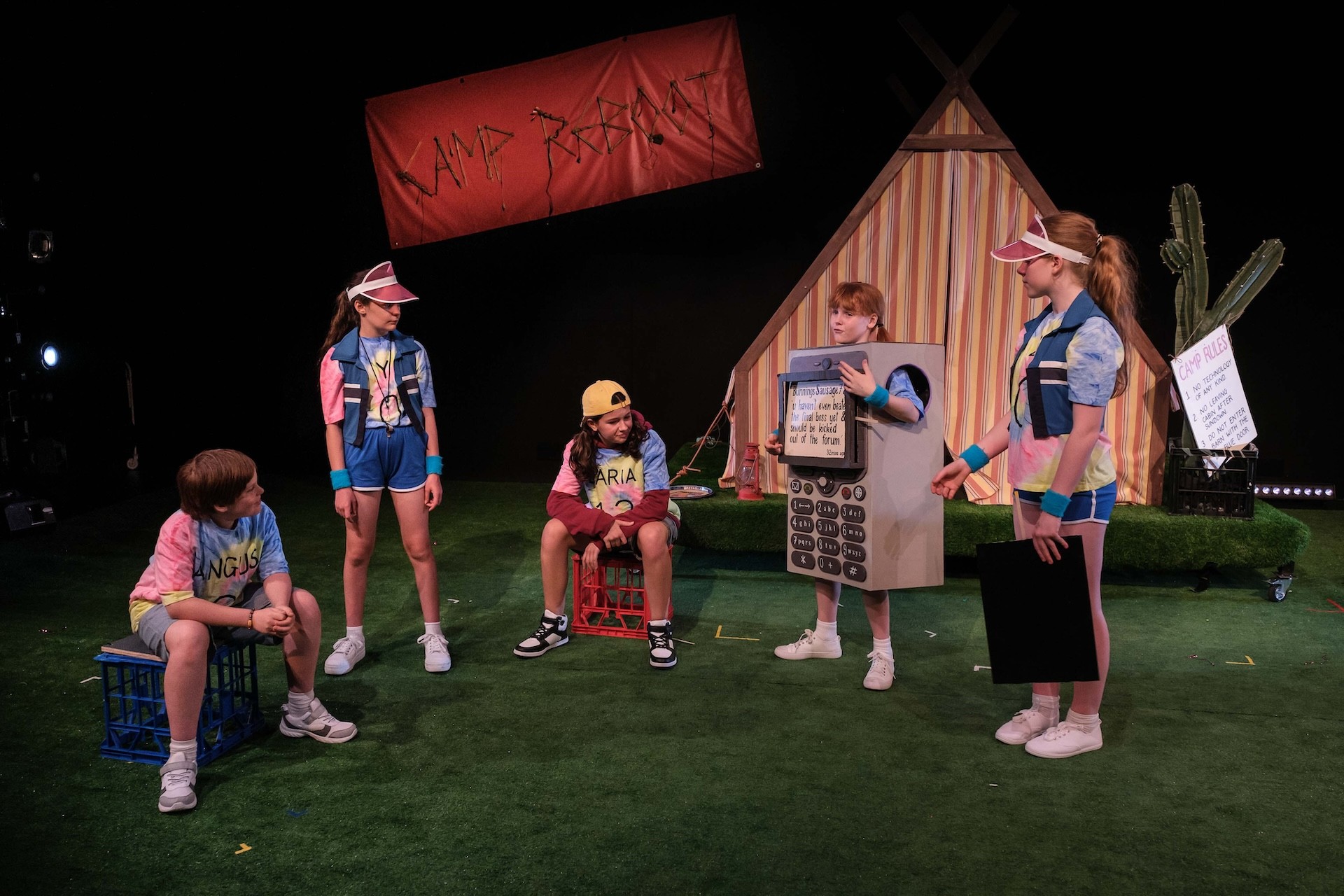 Five campers stand in a semicircle on the grass. One performer wears a large cardboard costume shaped like an oversized mobile phone, while others in bright tie-dye shirts react around them. A “Camp Reboot” banner hangs overhead.