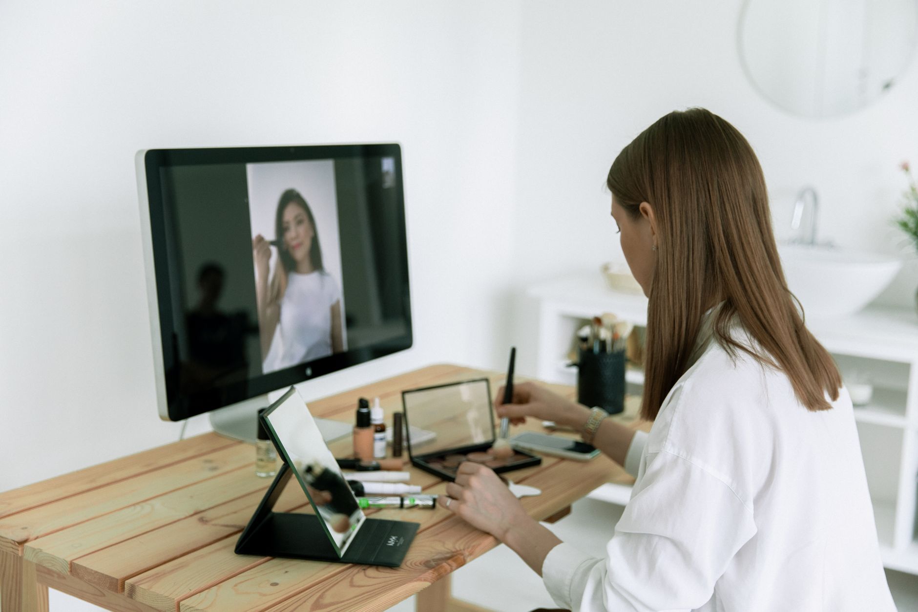 A woman follows an online makeup tutorial using a computer and tablet at home.