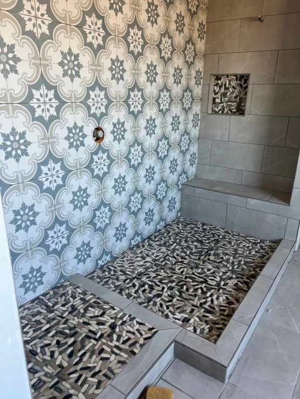 Modern shower tile installation in progress with decorative grey and white patterned wall tiles, coordinating grey large format tiles on a built-in bench, and unique multi-toned elongated pebble mosaic shower floor tiles.