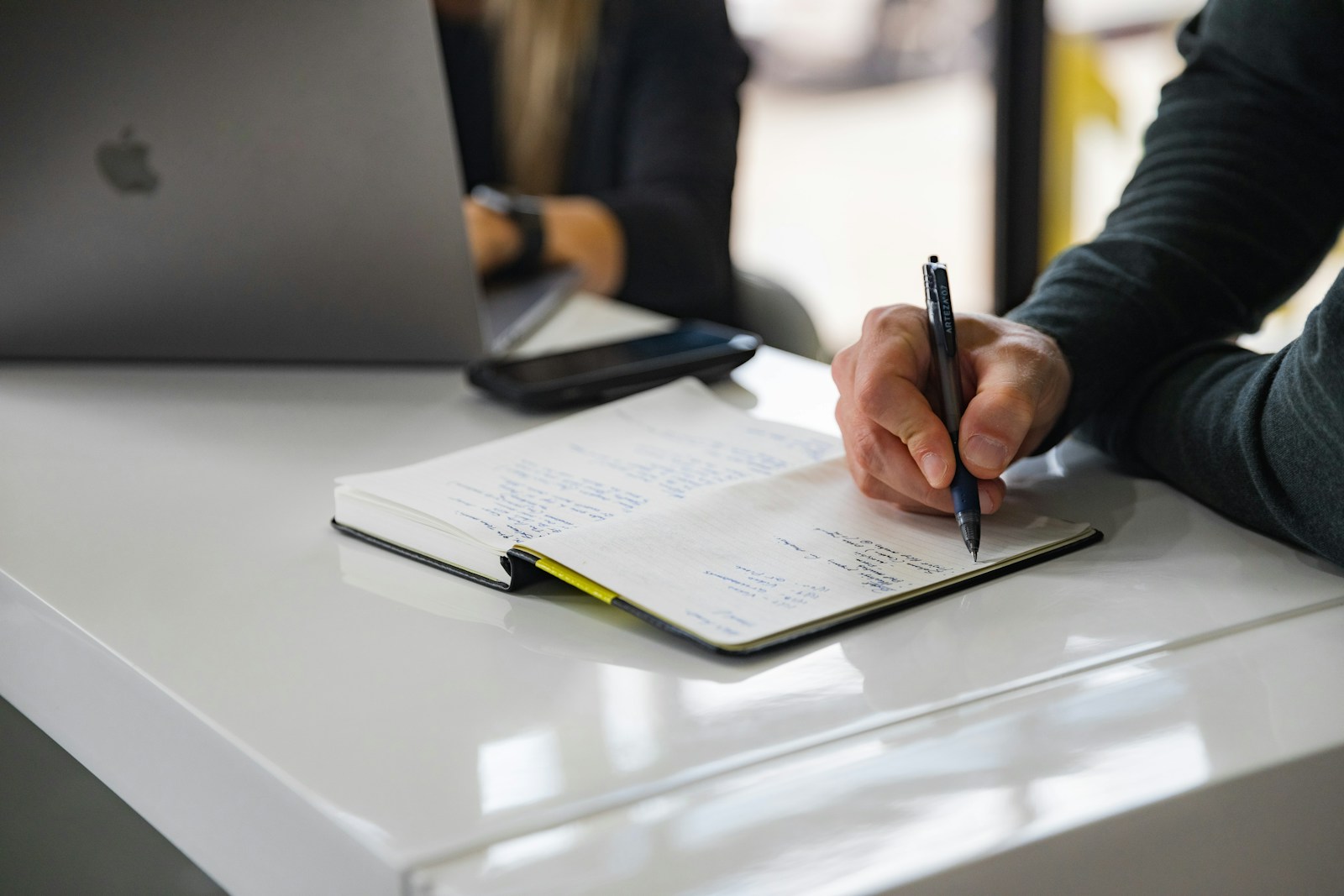 A person taking handwritten notes in a notebook at a desk with a laptop during a one-on-one nutrition consultation.