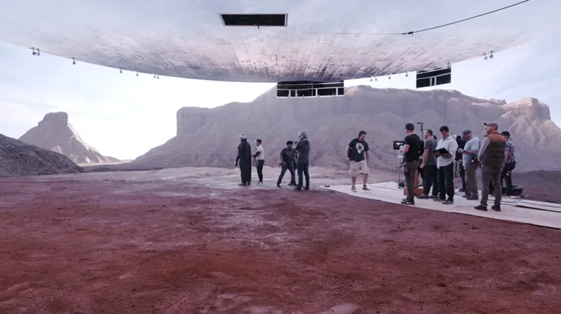 A group of people stands on a barren landscape under a large, hovering structure and dramatic skies.