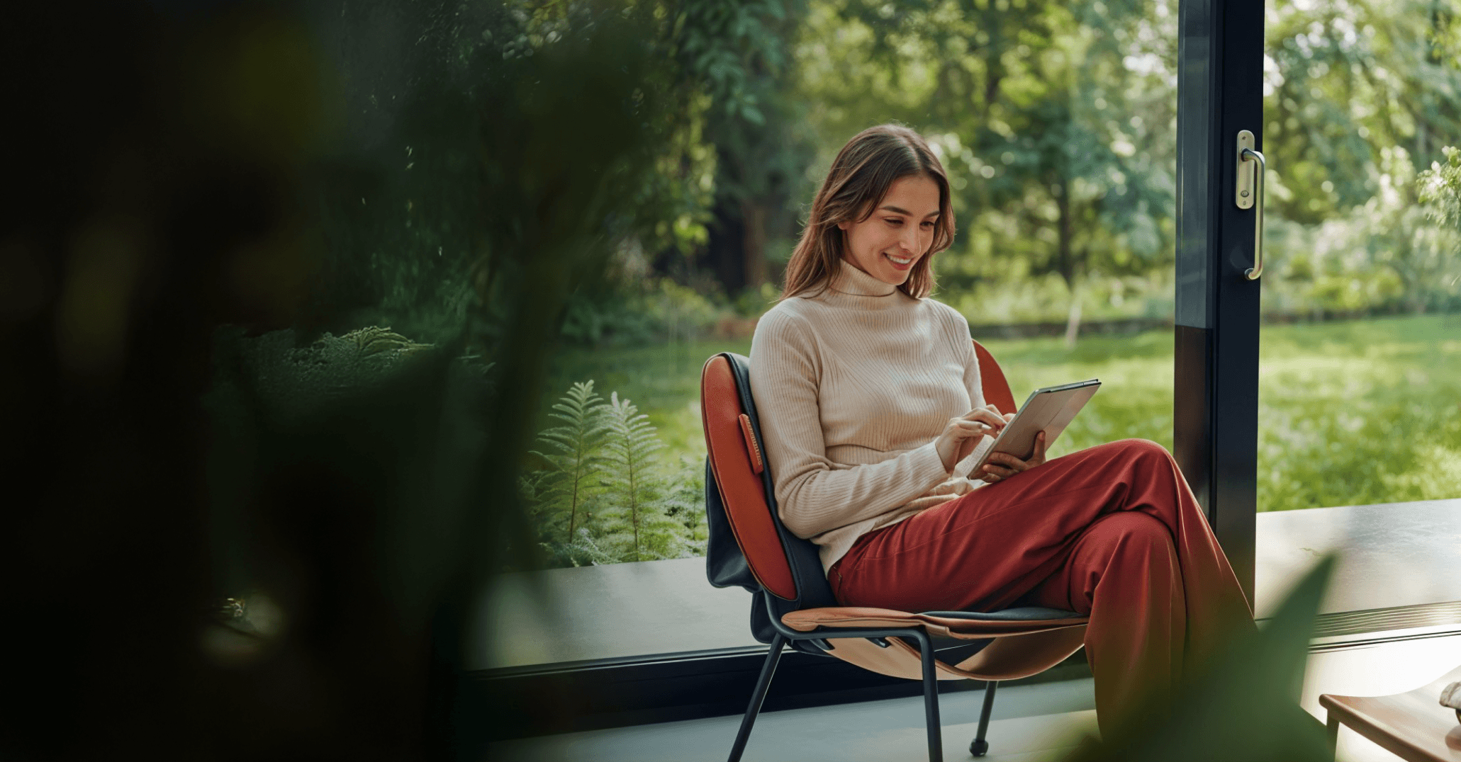 Check company name - Woman in turtleneck sweater and red pants smiles while looking at a tablet, sitting in a chair by a window.