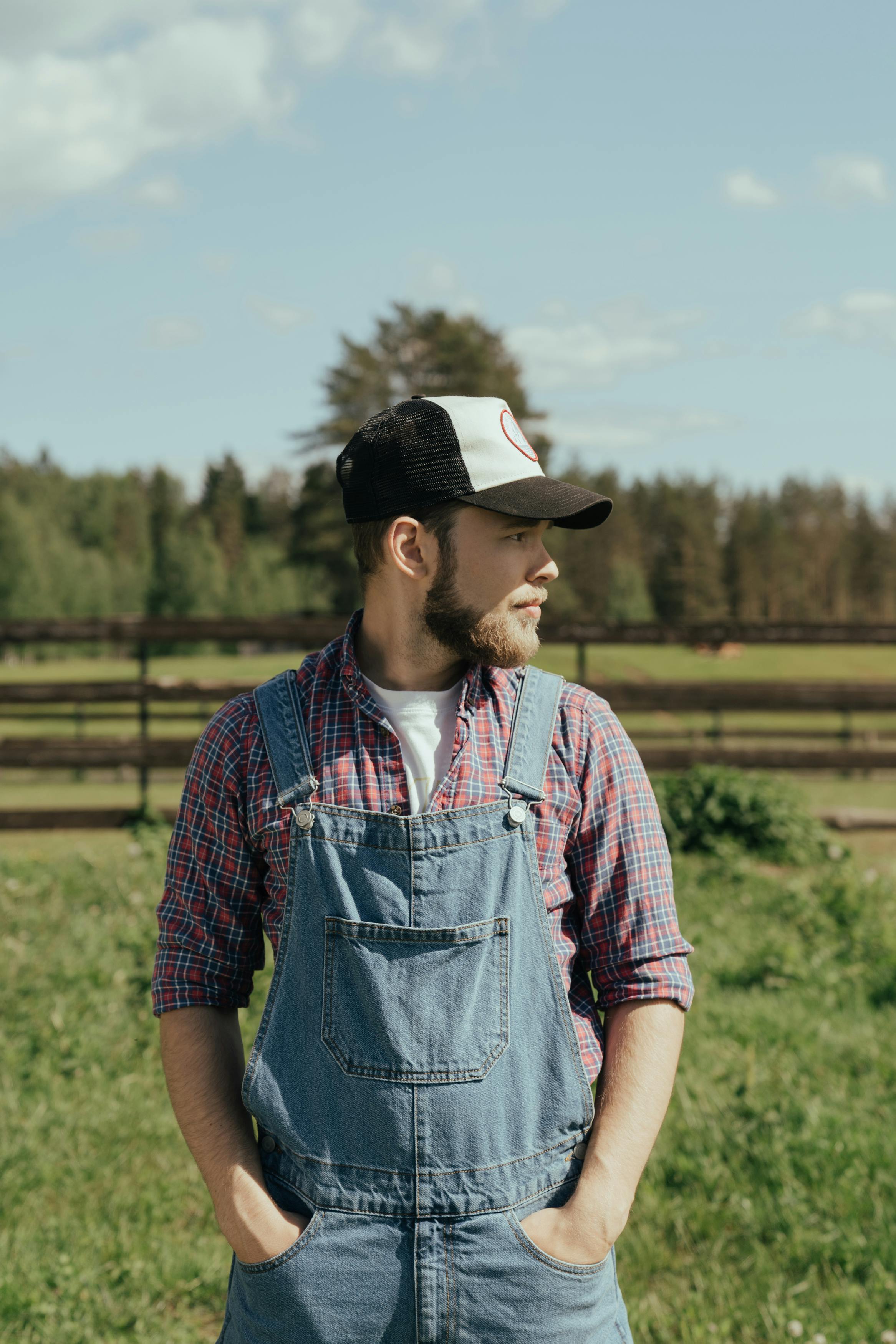 Bearded man in denim overalls, plaid shirt, and trucker hat stands in a sunny field with a wooden fence and forest.