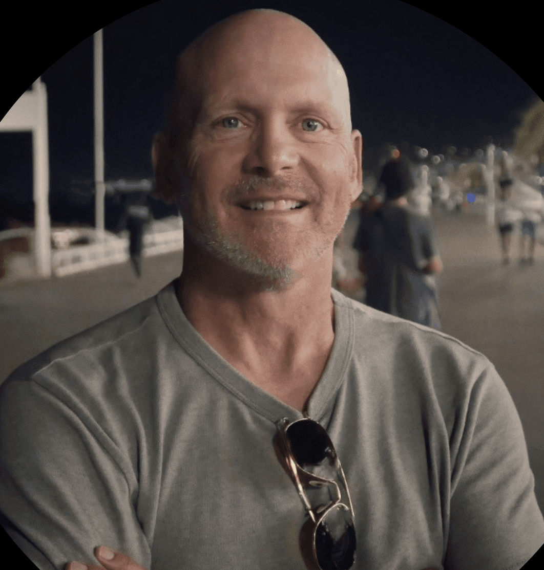 Director Jerry Brown, known for his work in public service announcements, comedy, and branding, smiles confidently while standing on a nighttime promenade. He wears a casual gray t-shirt with sunglasses hanging from the collar, giving off an approachable, laid-back vibe.