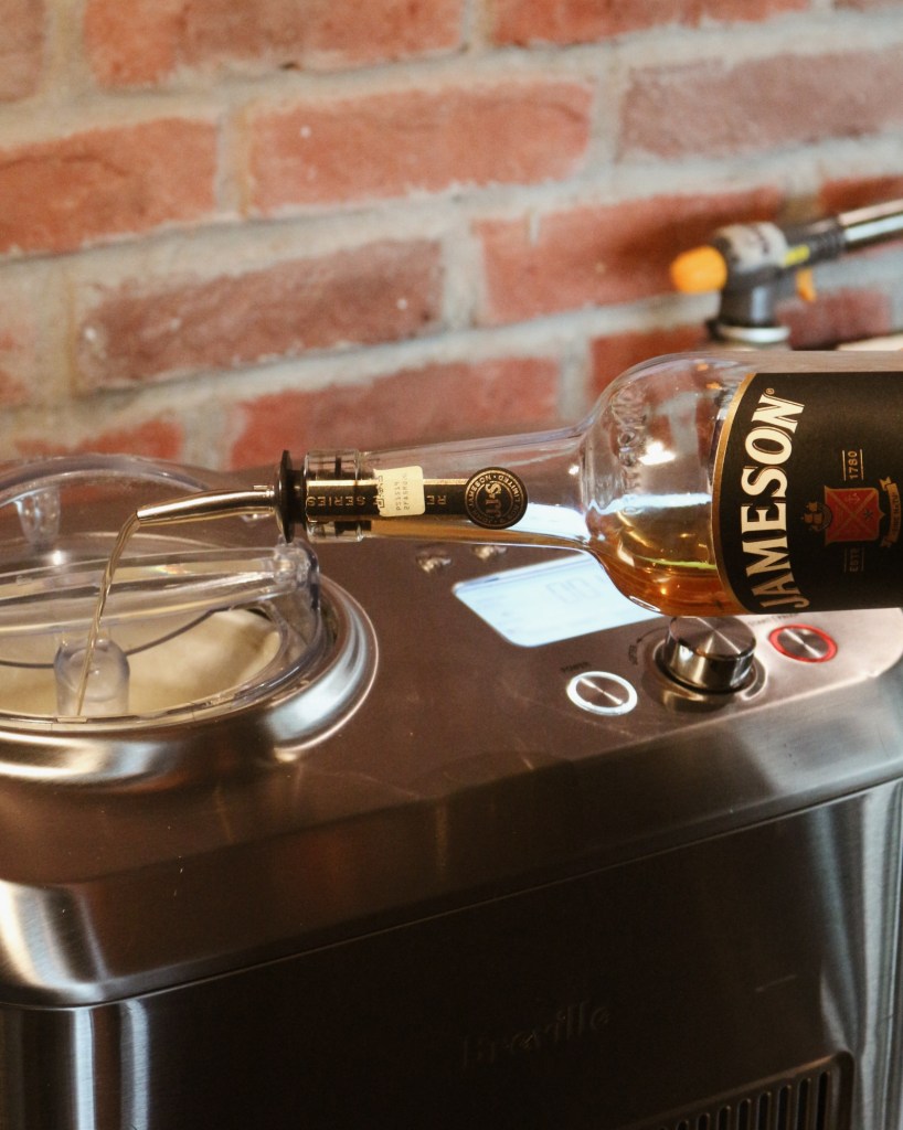 A Jameson whiskey bottle being poured into an ice cream maker, with a brick wall in the background.