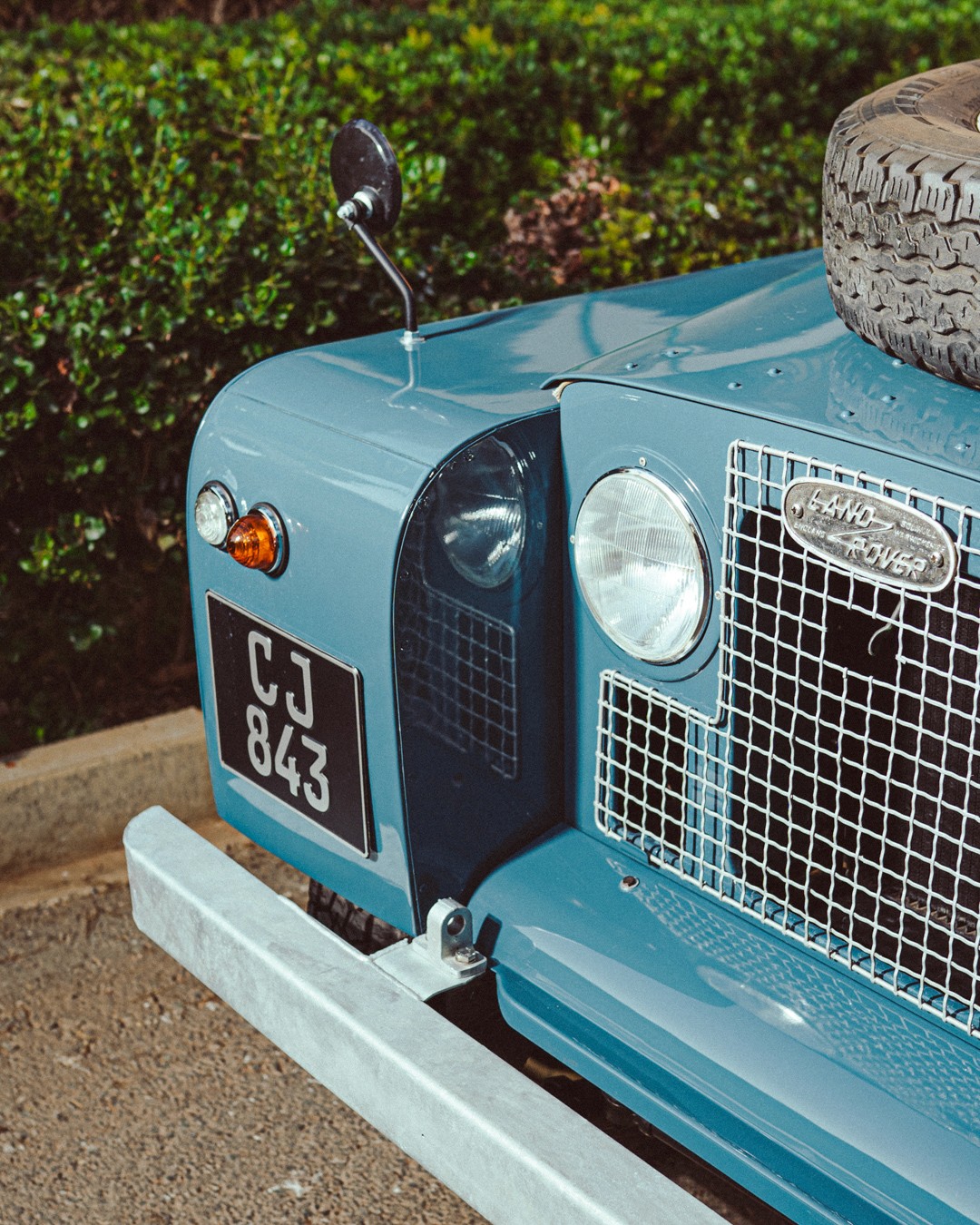 Front detail of a vintage blue Land Rover Series II, showing the mesh grille with "Land Rover" badge, round headlamp, indicator and a black number plate reading "CJ 843," with a hedge behind.