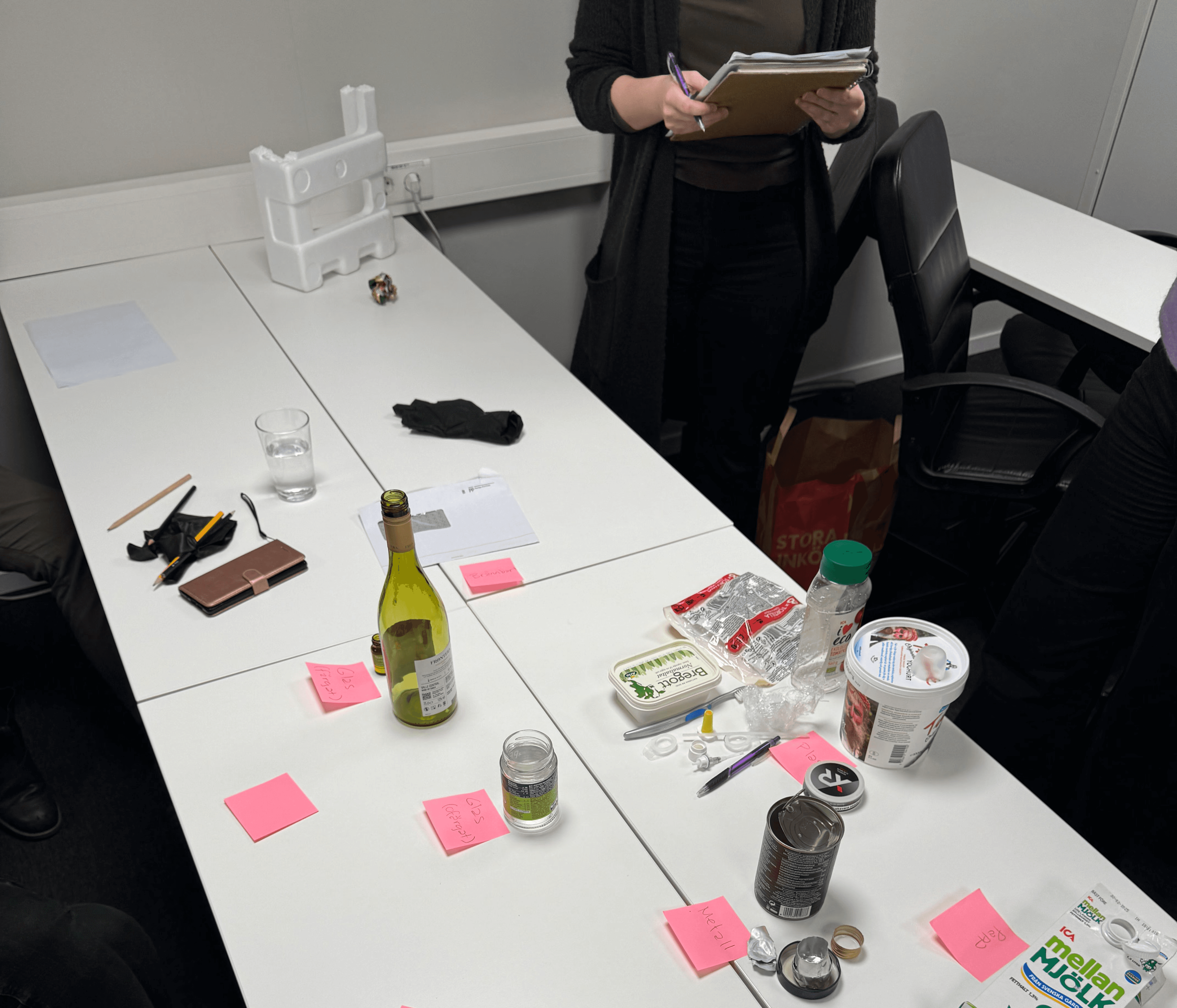 A recycling workshop in progress. A group is testing their ability to sort various waste items, including bottles, cans, plastic, and packaging, onto a table with labeled sticky notes. A facilitator is taking notes while participants engage in the sorting exercise.