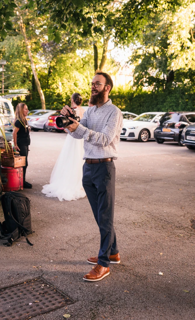 Tom Lally holding his camera in a car park while a bride in a white dress waits behind him