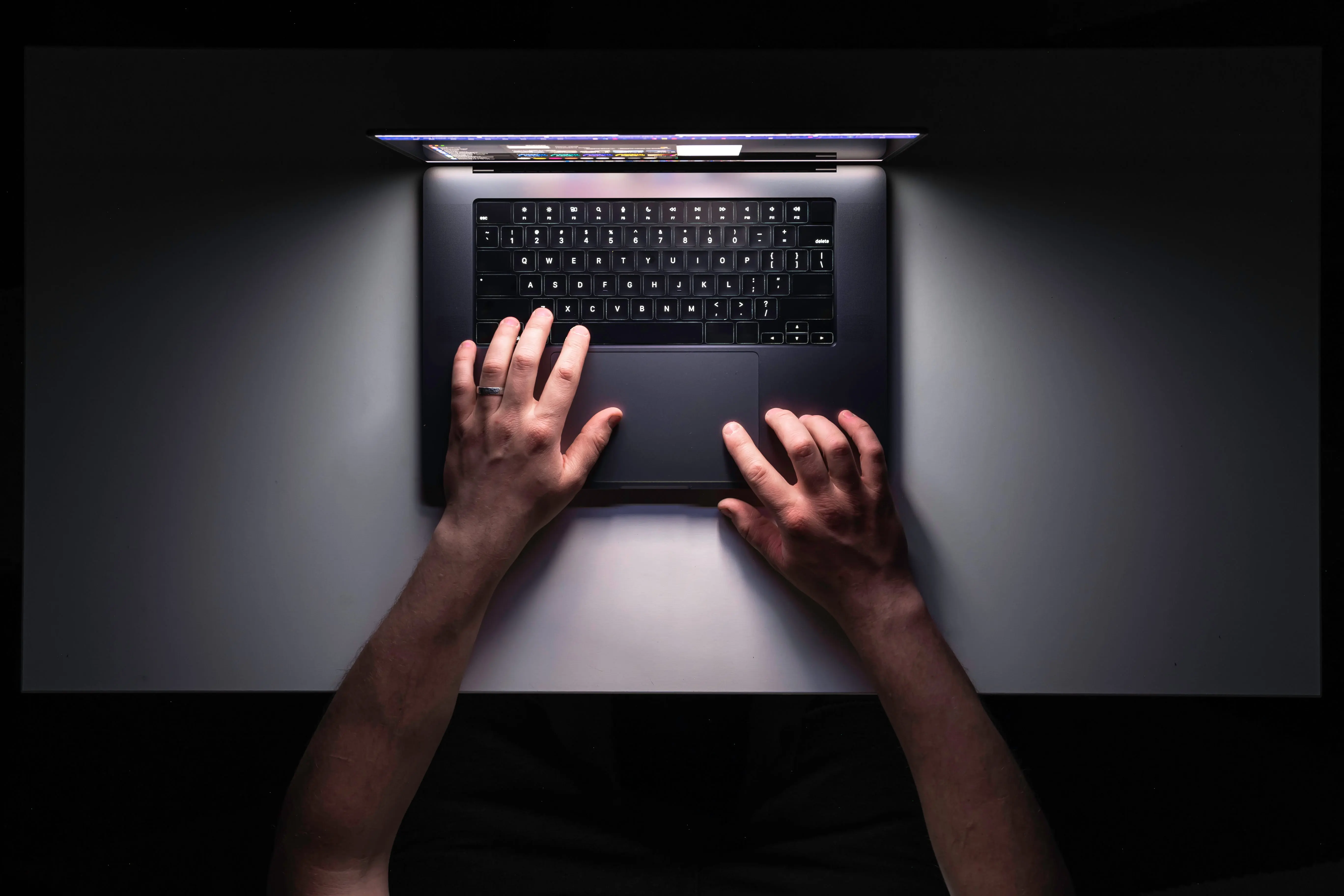 Top-down view of a person typing on a laptop in a dark setting, illuminated by the screen, highlighting hands on the keyboard and trackpad.