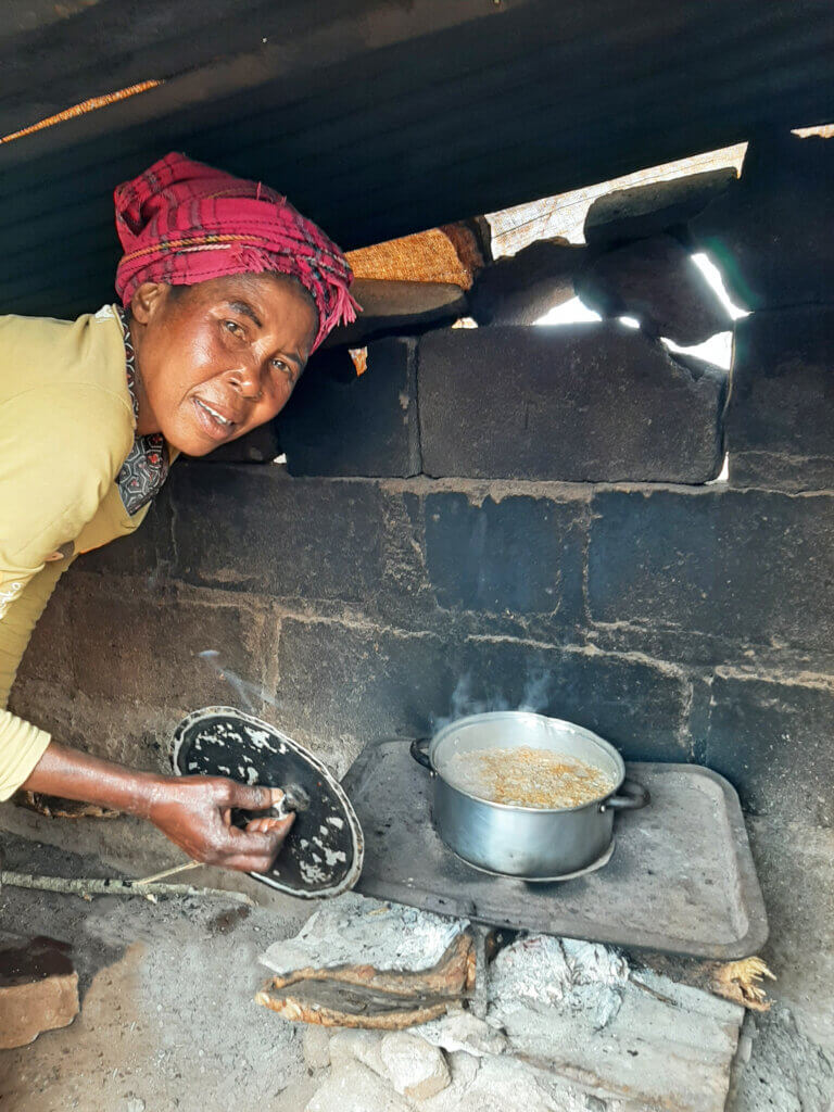 A woman lifts the lid of a pot cooking over an open fire inside a small stone and tin-roofed shelter.