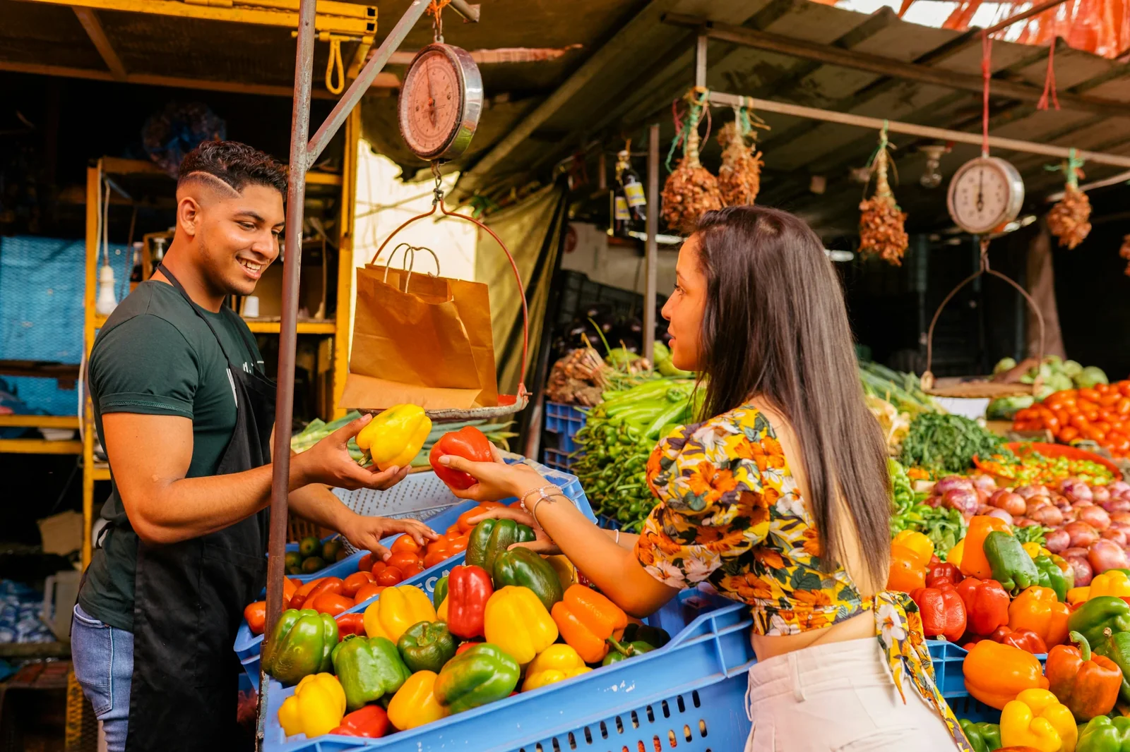 Young vendor and female customer exchanging colorful bell peppers at a vibrant outdoor market stall.