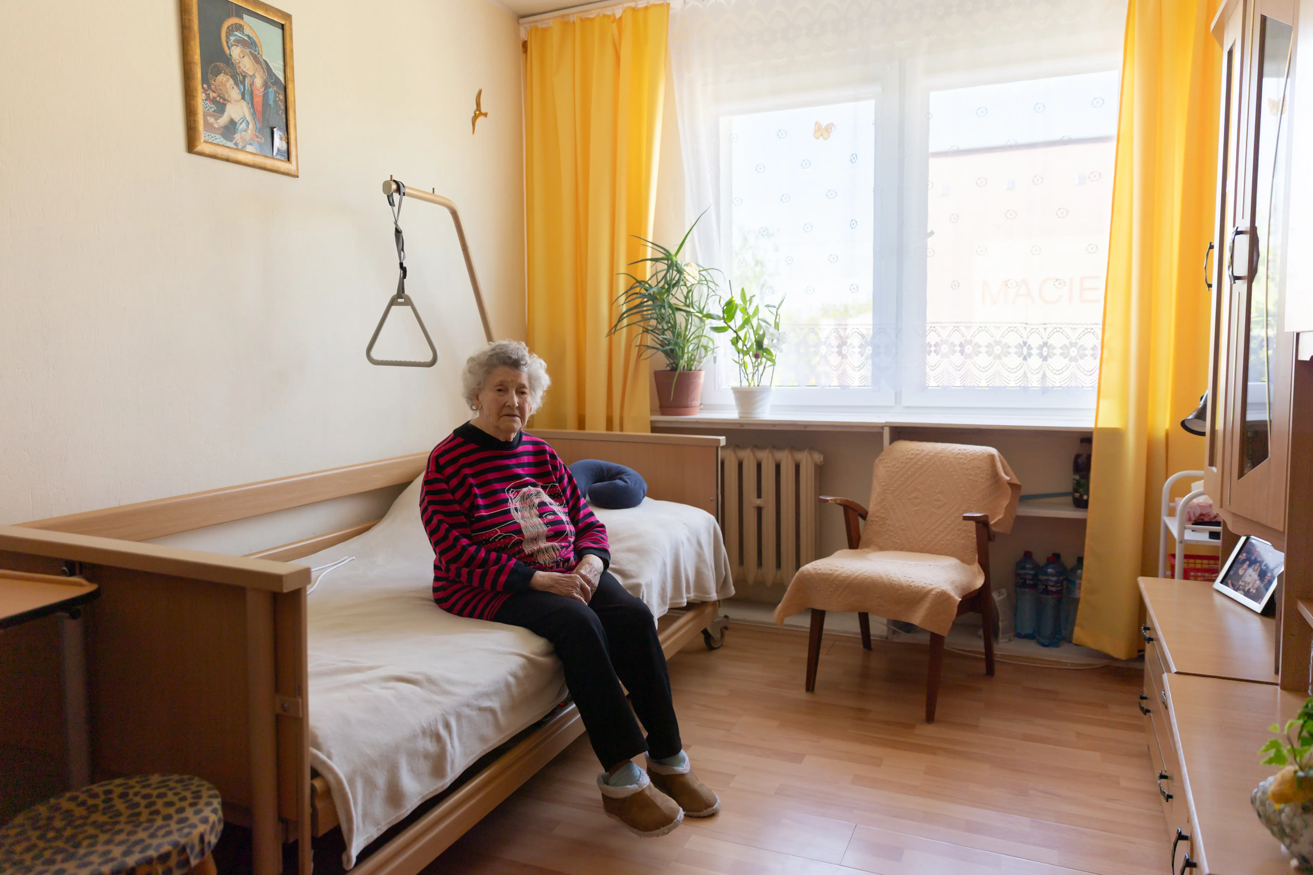 Starsza osoba siedzi na łóżku, nad którym wisi uchwyt do pomocy. Wokół sypialni z dużym, jasnym oknem.