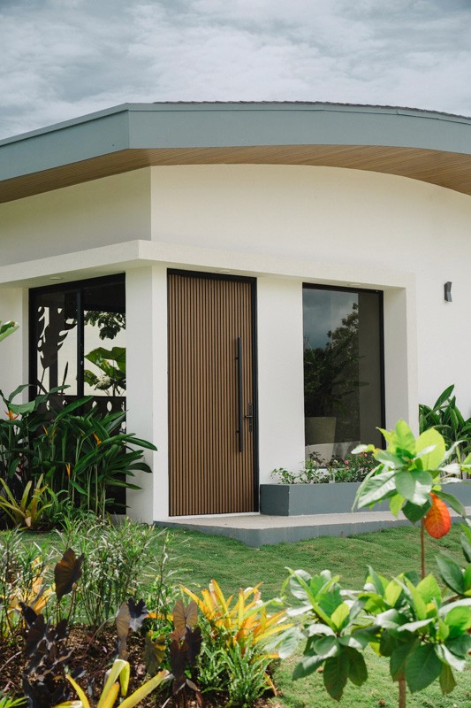 Modern rental villa exterior, wood-paneled front door, large windows, and lush tropical landscaping.