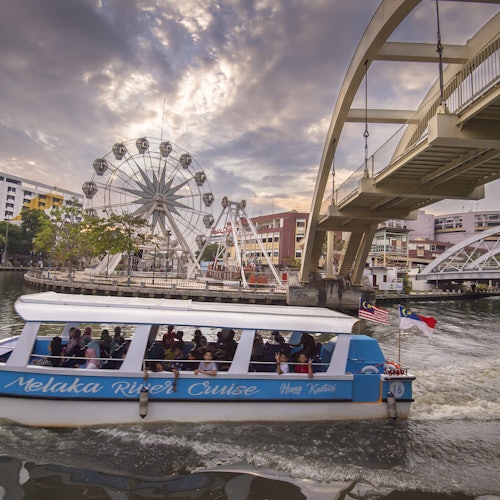 A boat with "Melaka River Cruise" written on it sails under a bridge; a ferris wheel and buildings are in the background.