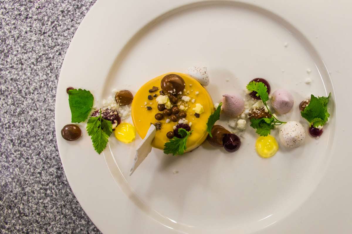 A top-down view shows a white plate featuring a yellow dessert topped with chocolate and nuts, garnished with mint leaves and berries.