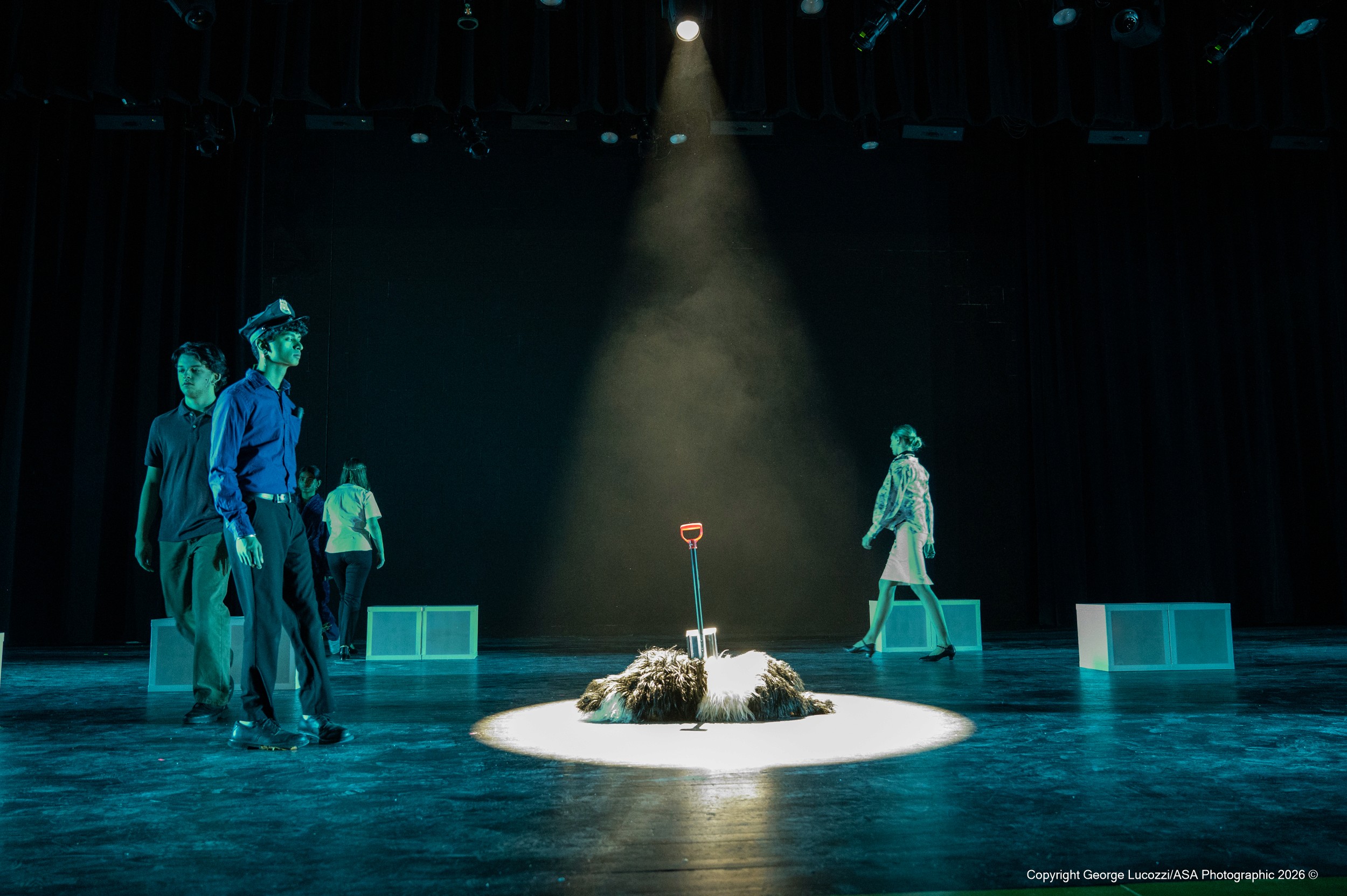 Center stage, Wellington—the black-and-white dog—lies motionless under a stark white overhead spotlight, a garden fork embedded in his body and oriented toward upstage. Surrounding him, ensemble members portraying the “Voices” circle the space in teal-blue sidelight, their partially shadowed forms contrasting the clinical isolation of the bright central special.