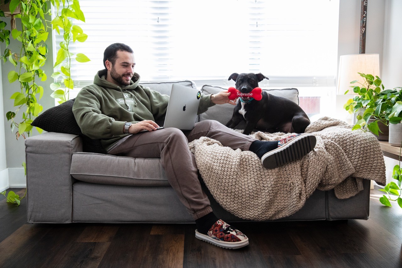 Wide lifestyle shot of a man relaxing on a couch with a laptop while his black dog plays beside him.