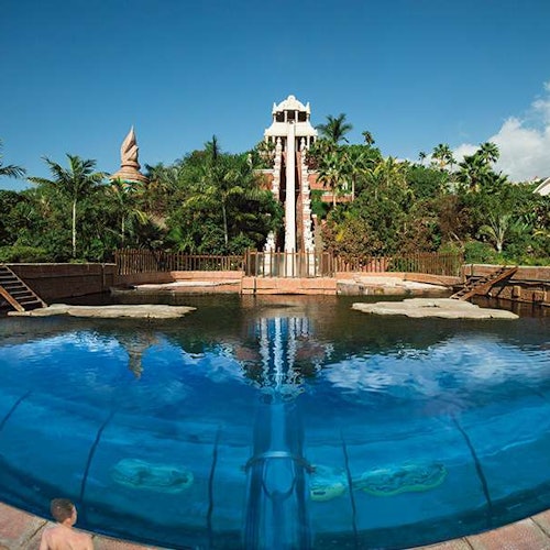 A large water slide resembling a temple ends into a pool, surrounded by foliage, with a child standing near the pool's edge.