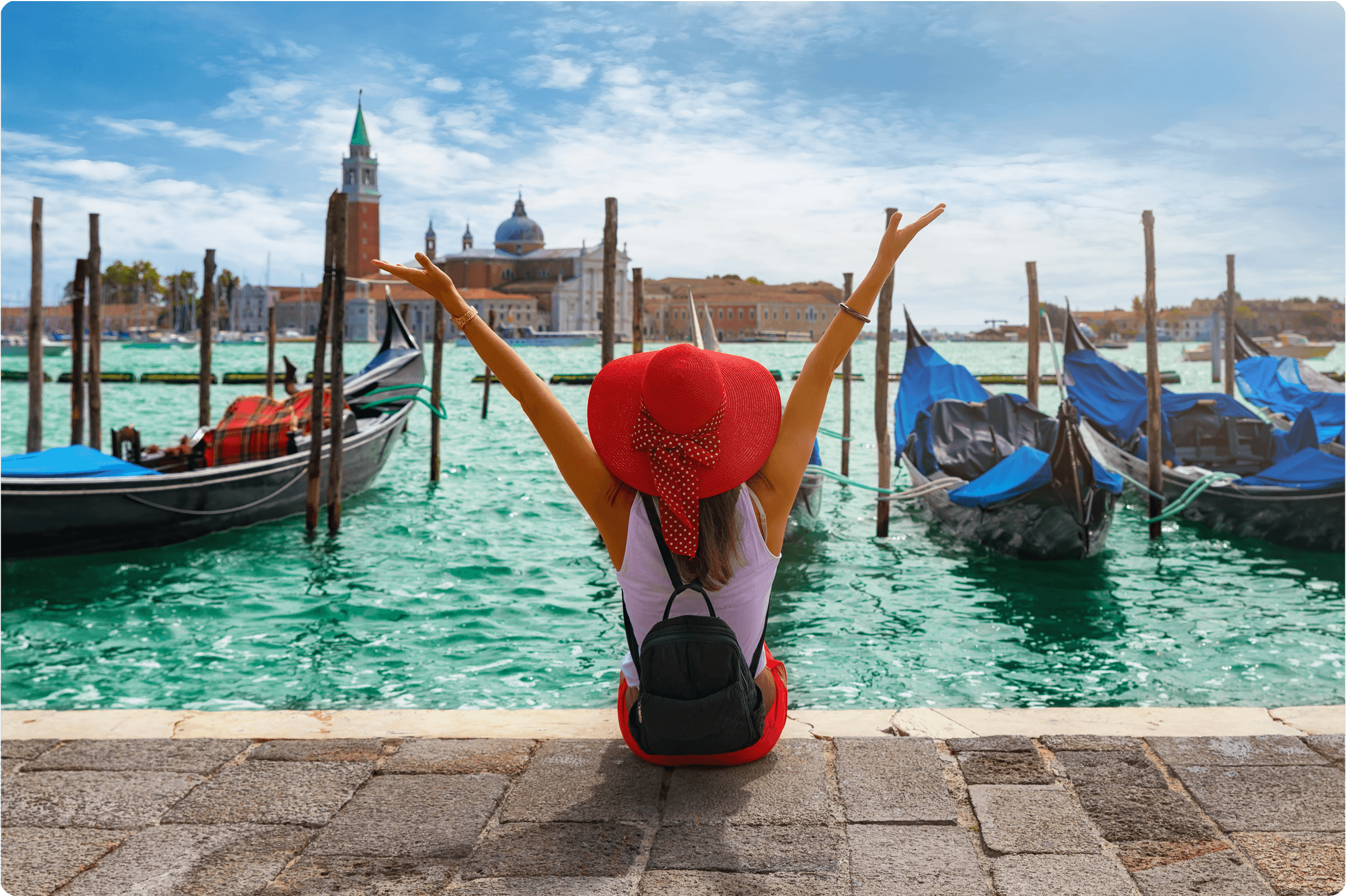 Woman in a red hat sitting by gondolas with arms raised on a sunny canal.