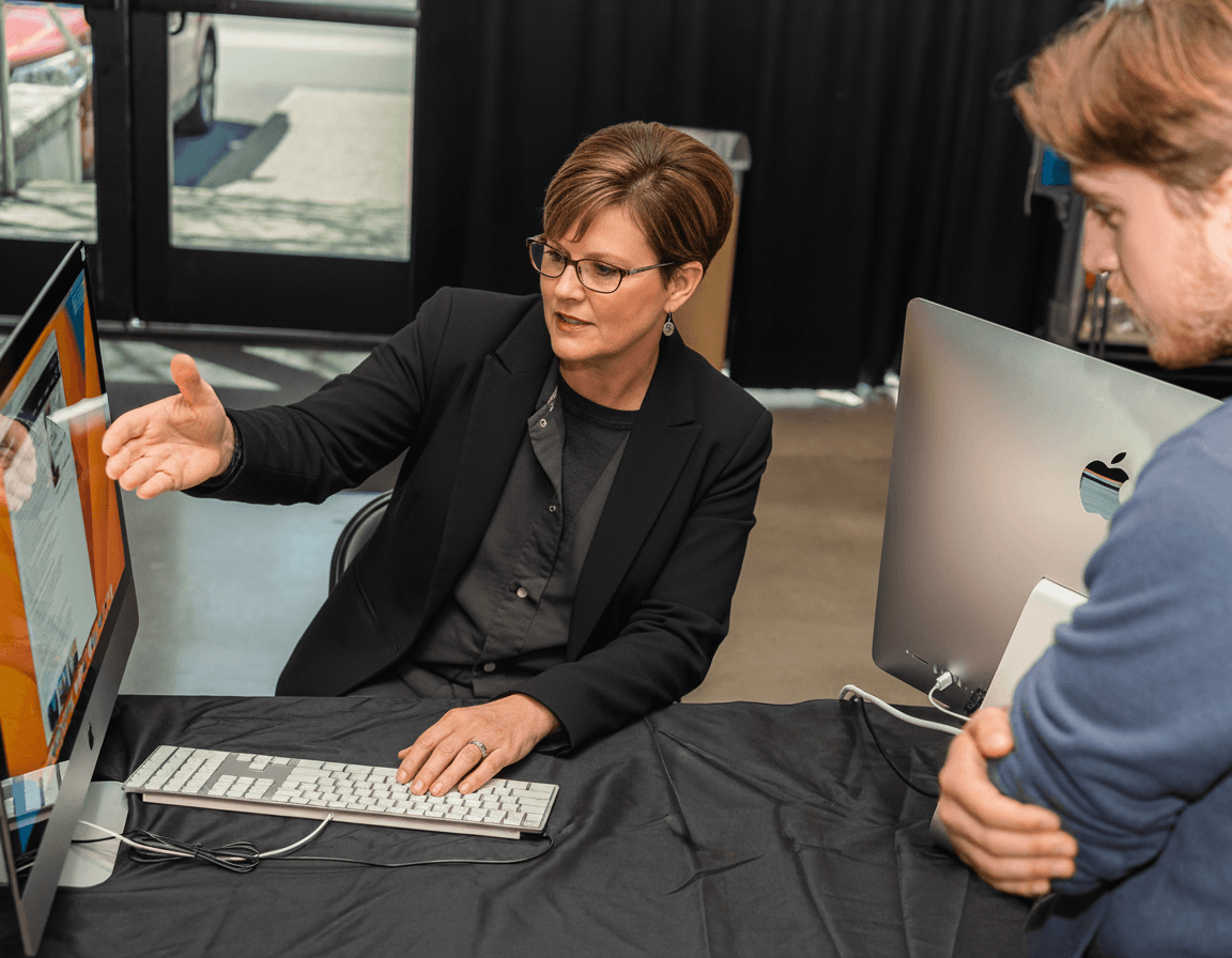 A woman in professional attire is seated at a desk, gesturing towards a computer screen displaying a presentation, while a man stands beside her observing attentively.