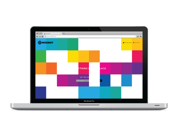 A silver MacBook Pro laptop is open, displaying a colorful, pixelated screen. The screen is divided into square blocks of varying colors, including blue, green, yellow, orange, pink, and purple. The text "MODBOT" is visible in the top left corner of the screen. Below it, the text "THING AND PLACE" is partially visible. The laptop’s web browser is open, showing a URL bar and address bar. The background is white.