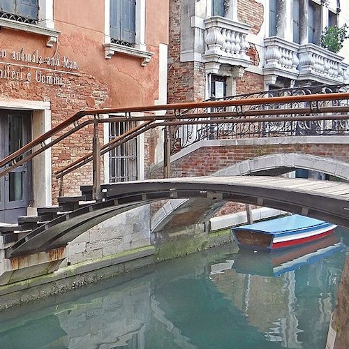 Un puente de madera arqueado se alza sobre un estrecho canal de Venecia, con una embarcación azul y roja amarrada en el agua bajo él.