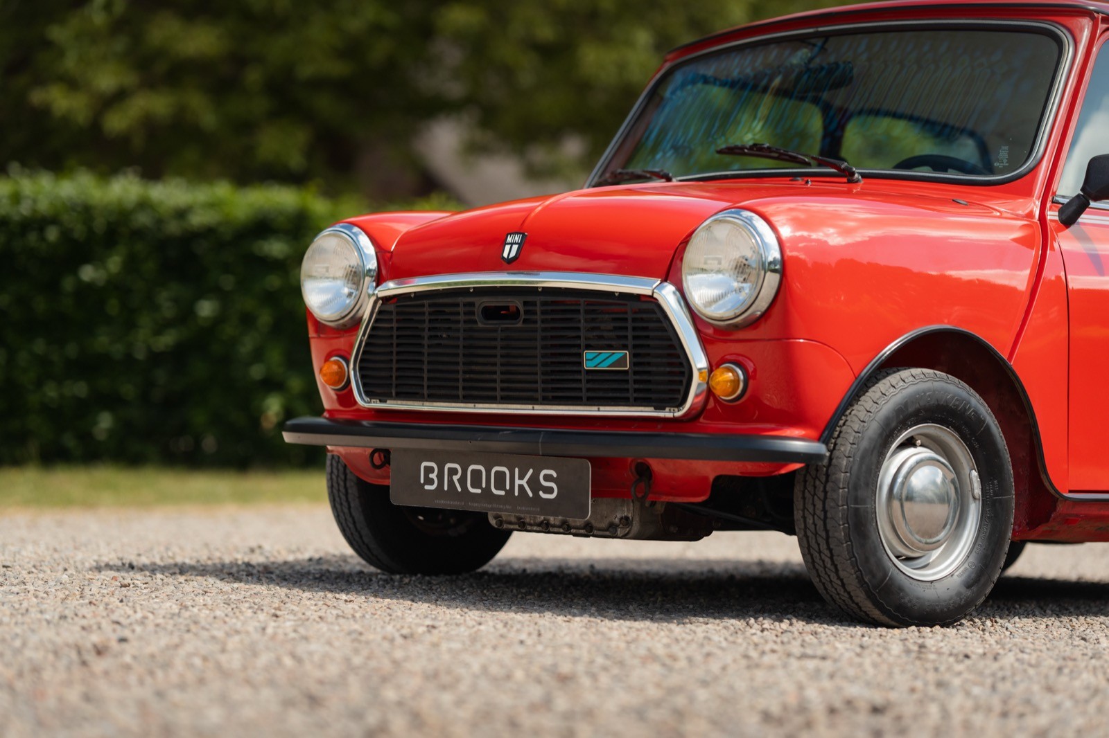 Classic red car parked on gravel, featuring a black grille and surrounded by green foliage.