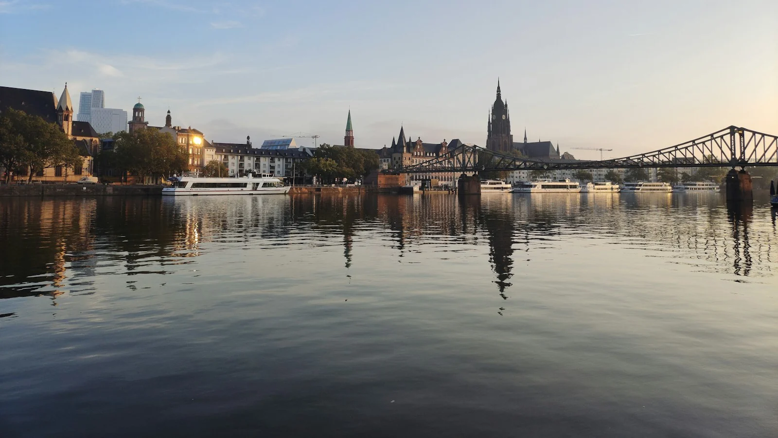 Der Eiserne Steg und der Frankfurter Dom im Morgenlicht, im ruhigen Main spiegeln sich das Ufer und die Ausflugsschiffe