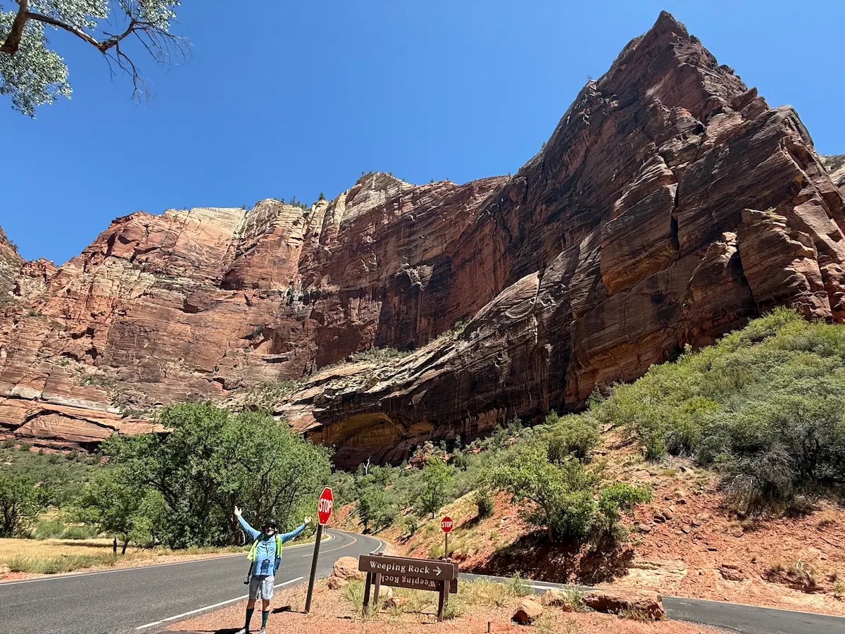 Stu standing with his arms raised in front of a massive rock formation near Weeping Rock in Zion National Park during a summer trip.