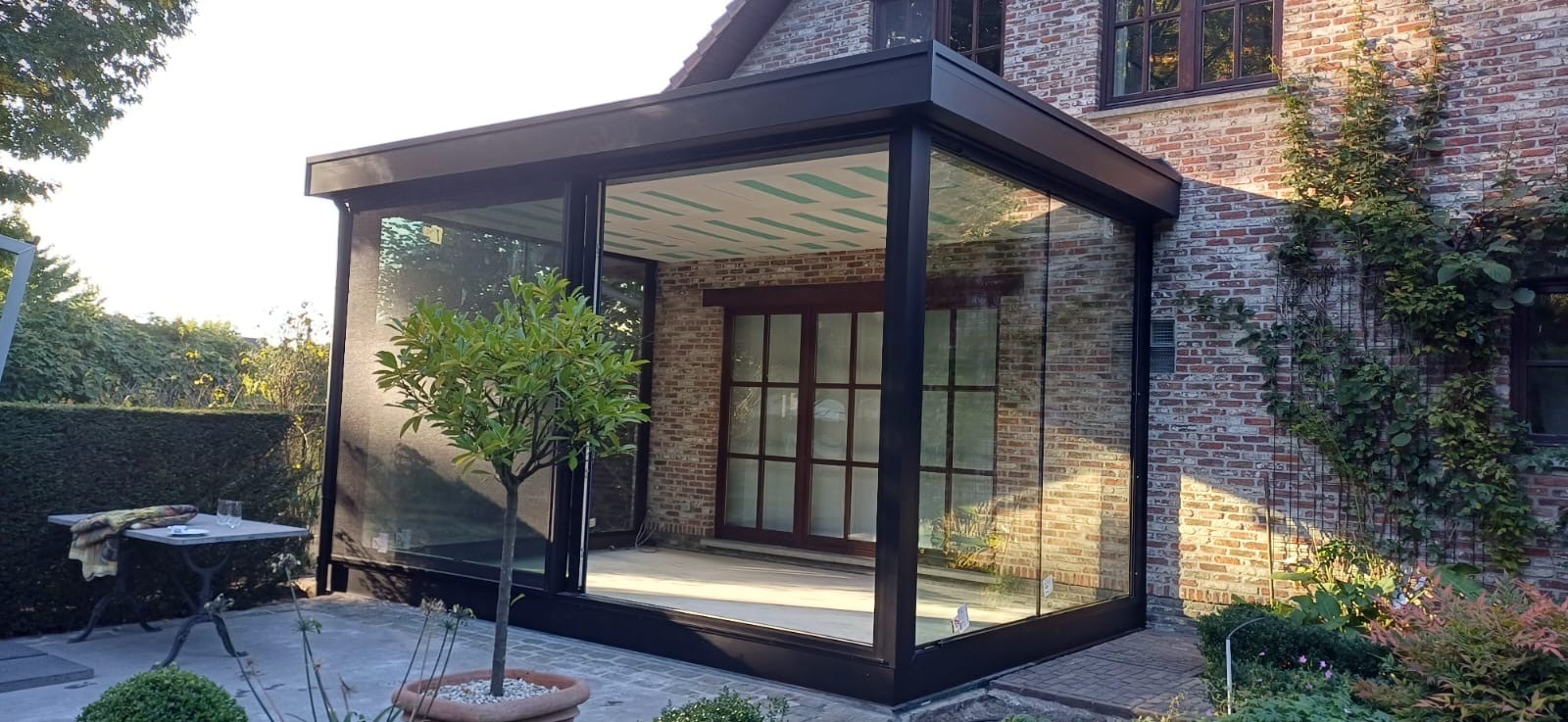 Buitenzicht op een moderne glazen aanbouw aan een bakstenen gebouw, met donker frame en een kleine boom in pot buiten. Het glas reflecteert de omgeving en de binnenkant van de aanbouw oogt licht en open.